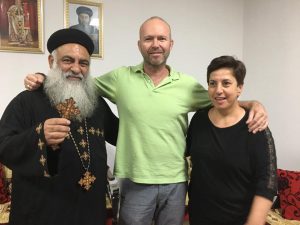 [Image description: Father Chris with Father Schnuder (left) and Onur (right). They are in a room with some framed icons behind them. Father Schnuder is holding up an ornate wooden cross. They are looking at the camera and smiling.]