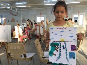 [Image description: a young girl holds up a painting she made. There are blue clouds in the sky, the sun is setting on the horizon. The focus of the painting is a long road disappearing into the horizon, in the middle of the paper, where a person is walking into the horizon carrying a bag and a walking stick. There are trees lining one side of the road and two houses on the other side of the road.]