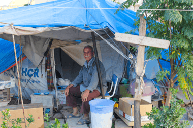 [Image description: another photograph of a tent. There is a man sitting under a tarp, outside his tent. There is a cross, made of two pieces of wood tied together, tied to one of the poles holding op the tarp.]