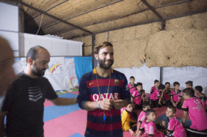 [Image description: Ninos Haddad, in a black shirt, and Allin, in a red-and-navy striped rugby shirt, stand in front of the camera but looking off to the side. Behind them, a group of boys sit on mats on a gym floor. The boys are wearing bright pink shirts.]