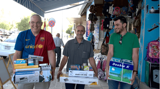 [Image description: Ashur and two other men are walking on the sidewalk outside the store, carrying boxes of paper, pens, a laminator machine, and other office supplies. They are all smiling.]