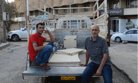 [Image description: Yusuf and Ashur sit on the bed of the truck, looking at the camera. There are a few boxes of supplies and more pieces of the desk in the bed of the truck.]