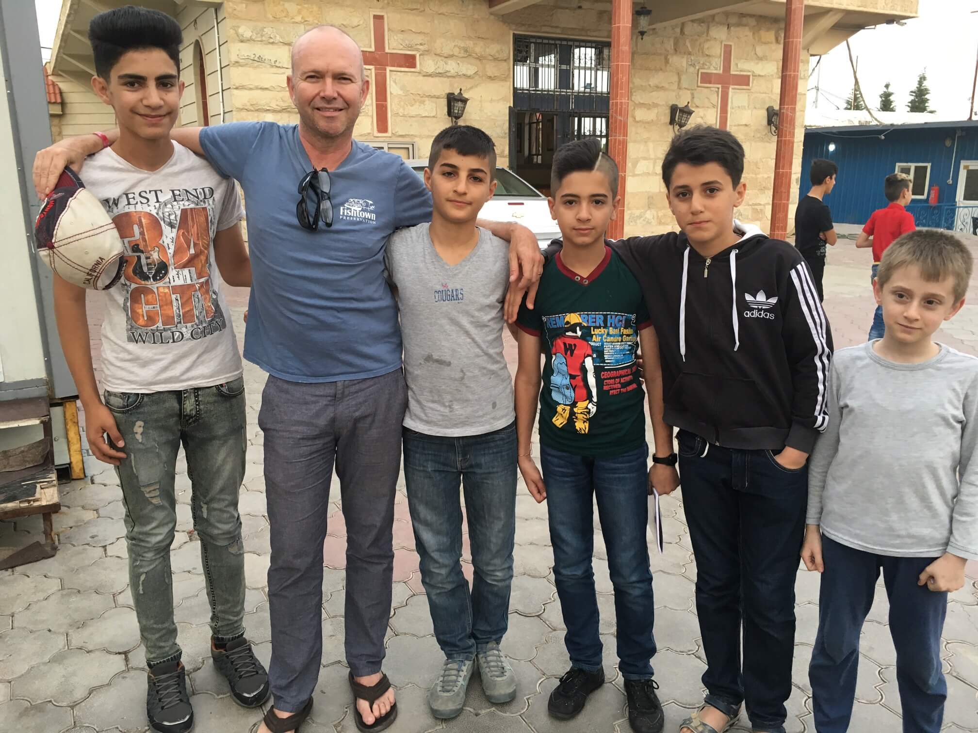 [Image description: Father Chris stands with five Iraqi boys in front of a building with crosses on the front. They are smiling, facing the camera, and have their arms around each other's shoulders.]
