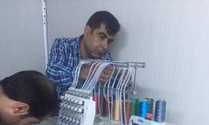 [Image description: A man arranges the threads on a sewing/embroidery machine. Another man is leaning over the maching, mostly off-camera, presumably setting up another part of the machine.]