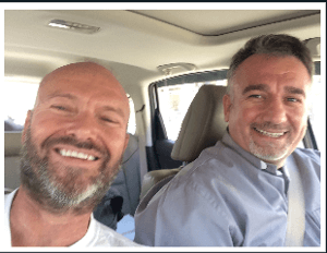 [Image description: selfie Fr. Chris, who has a beard, and Fr, Douglas Bazi. They are riding in a car, Fr. Bazi is in the driver's seat.]