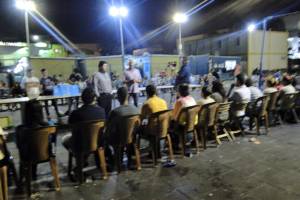 {Image description: photograph taken from behind a group of people sitting along the length of a long table. There are a few people standing in front of the table, talking to those sitting. It is night, they are gathered outside.]