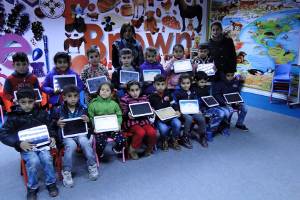 [Image description: elementary-aged students sit in two rows, facing he camera, displaying the electronic tablets donated to their school by SWIC, thanks to your generous donations.]
