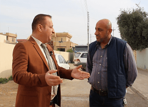 [Image description: two men stand outdoors, on a street or walkway, talking. The man on the left, in a brown suit jacket and blue tie, is speaking, with his hands out to his sides, palms up. The man on the right, in a blueish-purpliesh button-down shirt and sleeveless jacket, is looking at the man on the left and listening. He has is hands behind his back.]